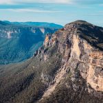 A breathtaking view of the Blue Mountains in New South Wales, Australia, with towering sandstone cliffs, lush forested valleys, and rolling hills under a clear blue sky.