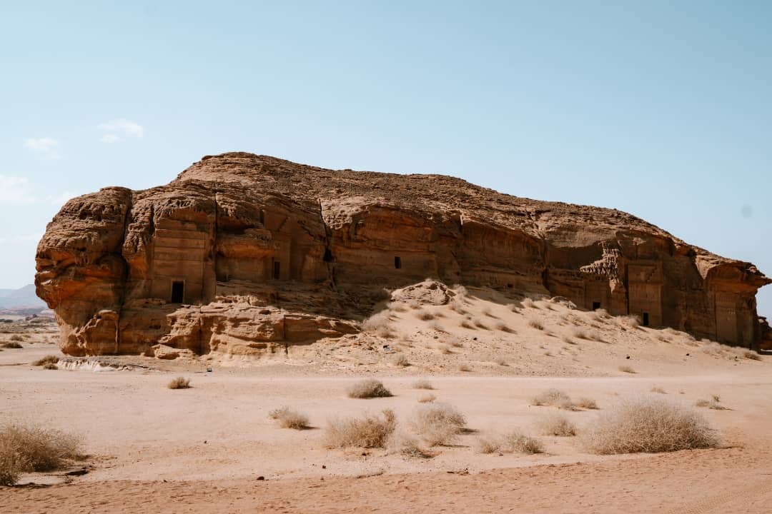 A view of the ancient rock-carved Hegra Burial Tombs in AlUla, Saudi Arabia, surrounded by a desert landscape with sparse vegetation under a clear blue sky. The intricate carvings on the sandstone hint at the historical significance of this UNESCO World Heritage Site.