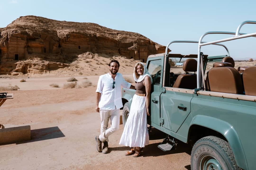 A couple poses beside a vintage green Land Rover during a tour of Hegra in AlUla, Saudi Arabia, with ancient rock-cut tombs in the background. The sunny desert setting highlights the charm of this UNESCO World Heritage Site.
