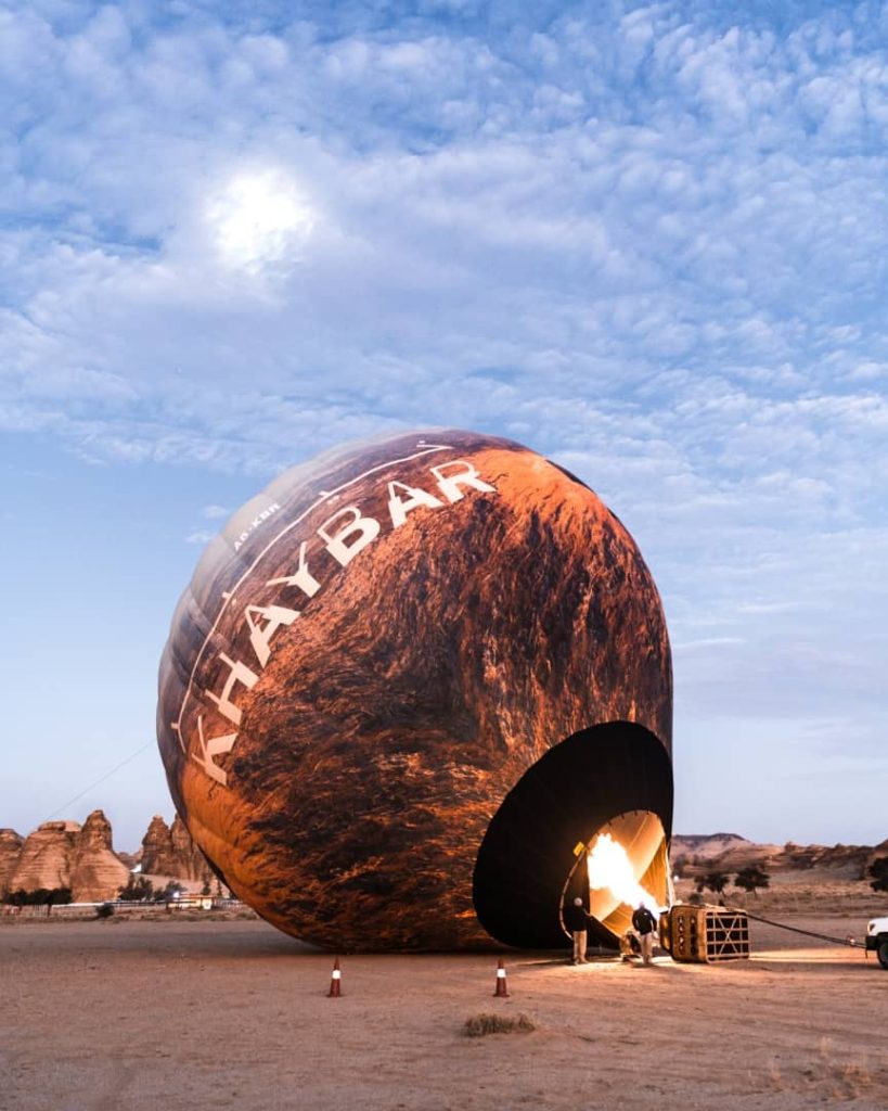 A hot air balloon being inflated in the desert of AlUla, Saudi Arabia, with the text "KHAYBAR" printed on its surface. The early morning sky is painted with soft clouds as the balloon prepares for a serene ascent.