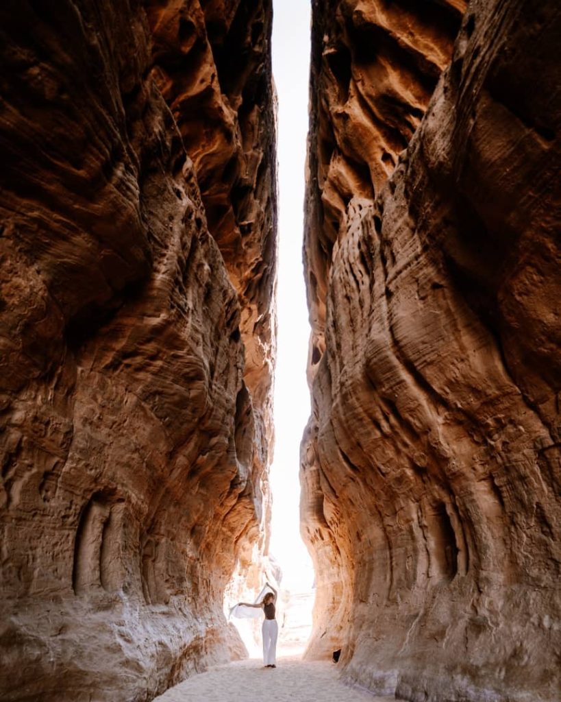 A woman stands in a narrow canyon at Jabal Ithlib in AlUla, Saudi Arabia, surrounded by towering sandstone walls with intricate natural textures. Light pours in from the open sky above, highlighting the dramatic formations and her flowing outfit.