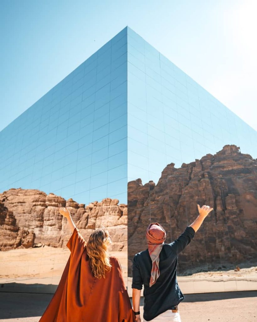 A couple stands in front of the Maraya Concert Hall in AlUla, Saudi Arabia, pointing towards its reflective mirrored surface that perfectly blends with the surrounding sandstone cliffs. The bright blue sky adds to the striking visual appeal of the modern structure.