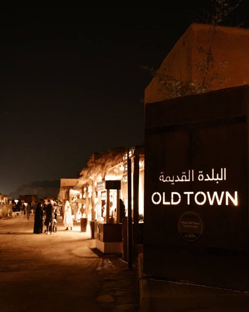 A night view of the Old Town in AlUla, Saudi Arabia, featuring an illuminated sign in Arabic and English that reads "Old Town." The warmly lit market stalls and people strolling along the pathways create a lively and inviting atmosphere.