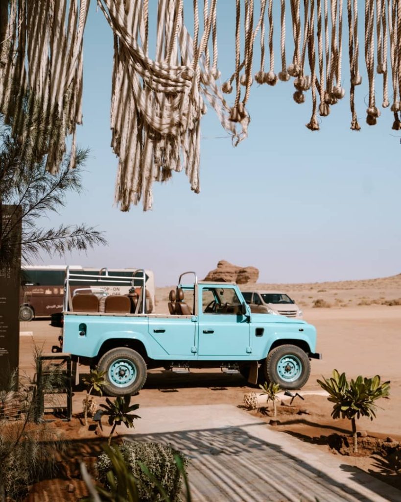A vintage turquoise Land Rover parked in a desert setting in AlUla, Saudi Arabia, framed by decorative hanging ropes and desert plants. The backdrop features sandstone formations and clear skies, highlighting the charm of this desert adventure.