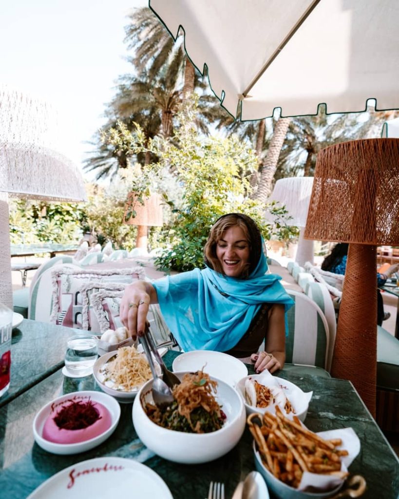 A woman draped in a blue scarf enjoys a vibrant meal at an outdoor restaurant surrounded by lush greenery and palm trees in AlUla, Saudi Arabia. The table is set with colorful dishes, including salads, fries, and dips, under a shaded umbrella.