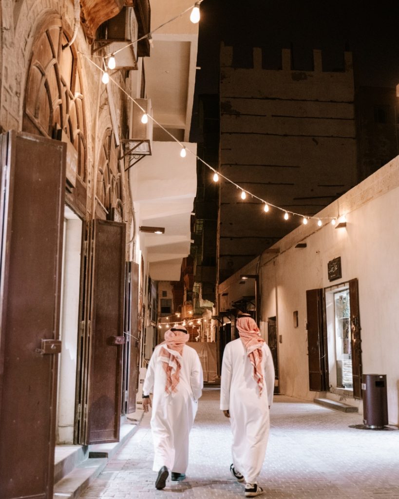 Two men in traditional Saudi attire walk through a narrow, well-lit alley in Jeddah’s historic Al-Balad district at night. String lights overhead and old-style architecture add a warm ambiance to the scene.