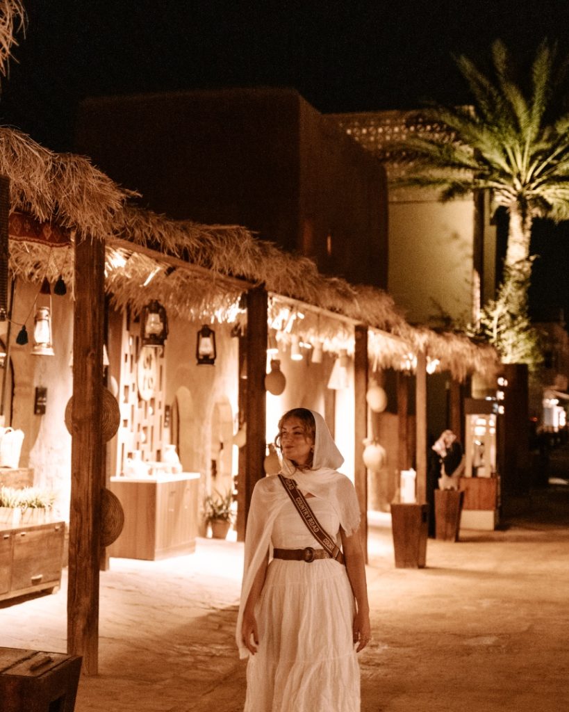 A woman dressed in a white flowing outfit strolls through the warmly lit market streets of AlUla's Old Town at night. The stalls, adorned with traditional decorations and palm-thatched roofs, create a serene and historic atmosphere.