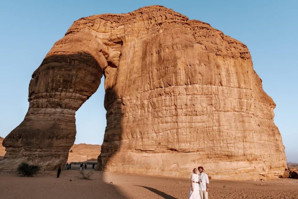 A couple dressed in white poses in front of the towering Elephant Rock in AlUla, Saudi Arabia, as the golden light of sunset illuminates the sandstone formation. The serene desert landscape surrounds the iconic natural landmark.
