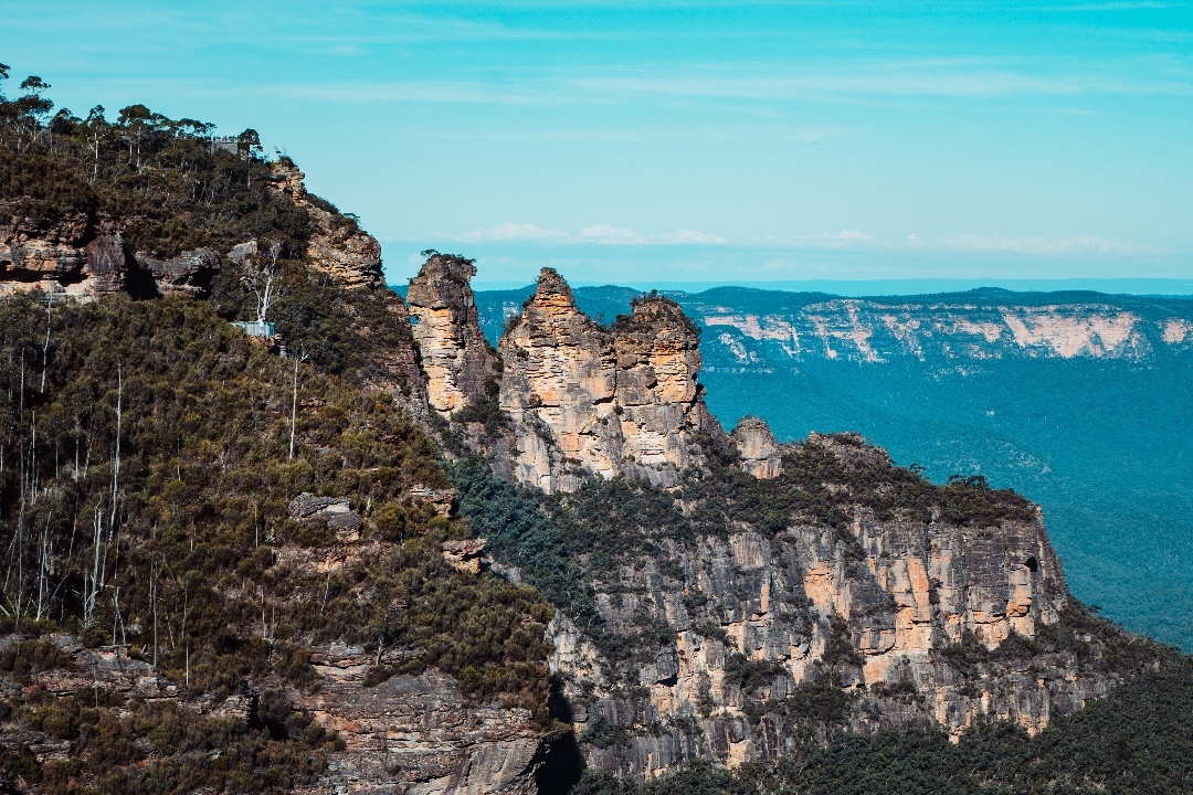 The iconic Three Sisters rock formation in the Blue Mountains, New South Wales, Australia, surrounded by lush eucalyptus forest and dramatic cliffs under a bright blue sky.