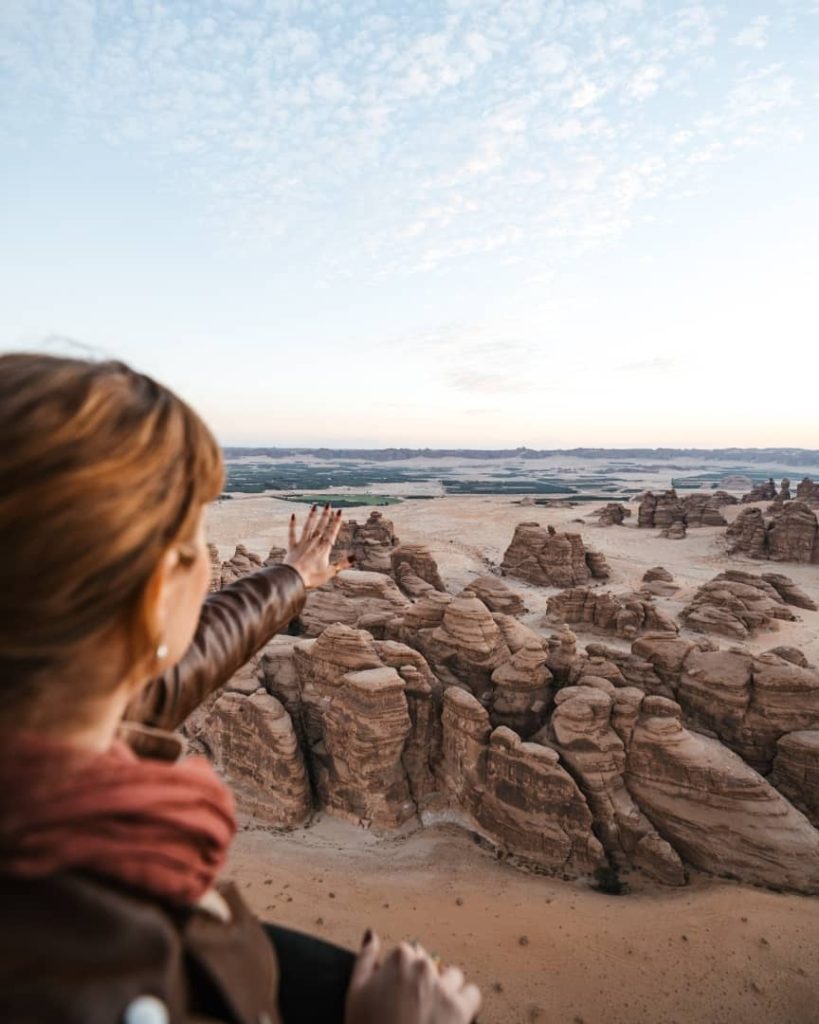 A woman extends her hand toward the dramatic desert landscape of Hegra, AlUla, Saudi Arabia, viewed from a high vantage point. The scene features unique sandstone formations set against a sprawling horizon under a sky filled with soft clouds.
