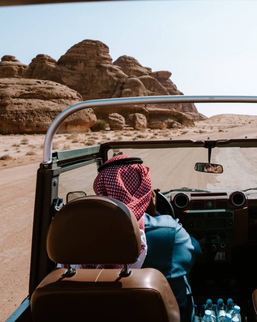 View from inside a vintage open-top vehicle driving through the desert landscape of Hegra in AlUla, Saudi Arabia. A driver wearing a traditional red-and-white shemagh navigates the sandy road surrounded by unique sandstone rock formations.
