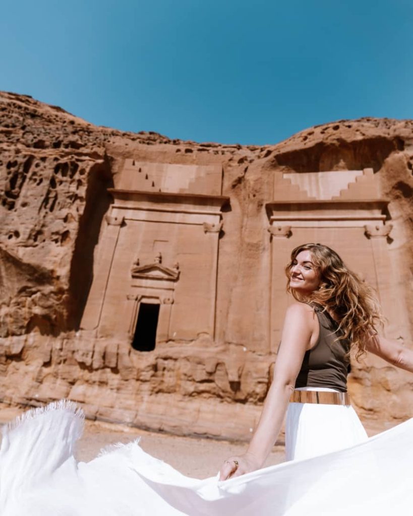 A smiling woman in a flowing white skirt poses gracefully in front of an ancient rock-cut tomb at Hegra, AlUla, Saudi Arabia. The tomb's intricate carvings contrast against the clear blue sky, emphasizing the site's historical grandeur.