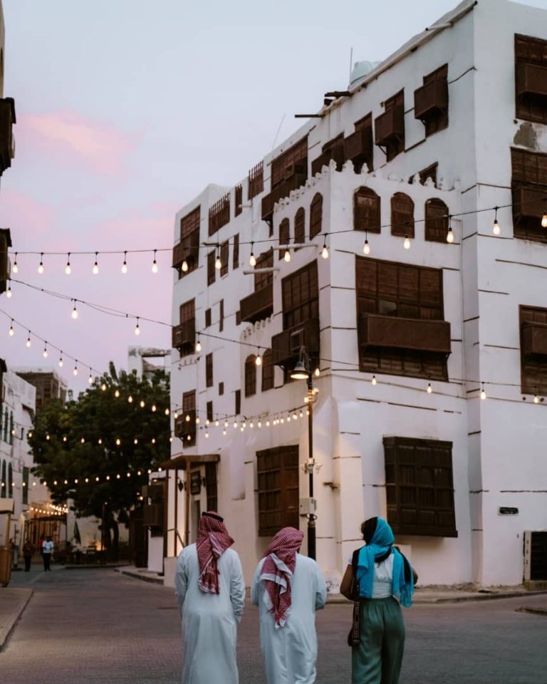 A charming street in Al-Balad, the historic center of Jeddah, Saudi Arabia, at dusk. The scene features traditional white buildings with wooden lattice windows, strung with glowing fairy lights. In the foreground, three men in white thobes and red checkered shemaghs, along with a woman wearing a blue scarf, walk down the cobblestone street.