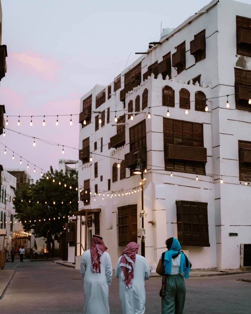 A charming street in Al-Balad, the historic center of Jeddah, Saudi Arabia, at dusk. The scene features traditional white buildings with wooden lattice windows, strung with glowing fairy lights. In the foreground, three men in white thobes and red checkered shemaghs, along with a woman wearing a blue scarf, walk down the cobblestone street.