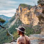 A man wearing a wide-brimmed hat relaxes at the edge of a natural infinity pool at Fortress Falls in the Blue Mountains, NSW. The view stretches out to towering sandstone cliffs and a deep green valley below. The golden hues of the rock formations contrast against the lush forest, creating a breathtaking scene.