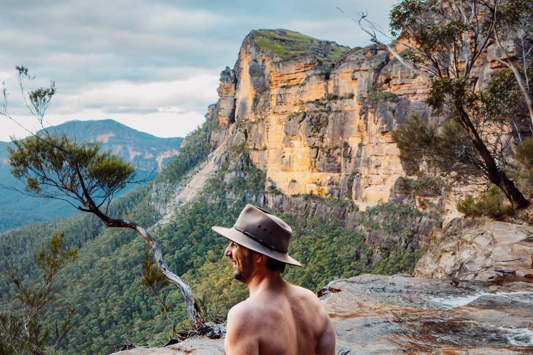 A man wearing a wide-brimmed hat relaxes at the edge of a natural infinity pool at Fortress Falls in the Blue Mountains, NSW. The view stretches out to towering sandstone cliffs and a deep green valley below. The golden hues of the rock formations contrast against the lush forest, creating a breathtaking scene.