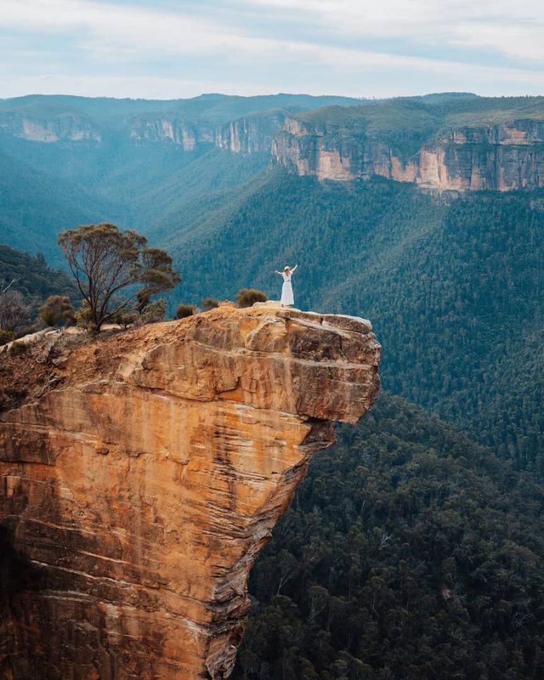 A person in a white dress stands with arms raised on the edge of Hanging Rock, a dramatic sandstone cliff jutting out over a dense forest in the Blue Mountains, NSW, Australia. The expansive landscape of rugged cliffs and green valleys stretches into the distance under a cloudy sky.