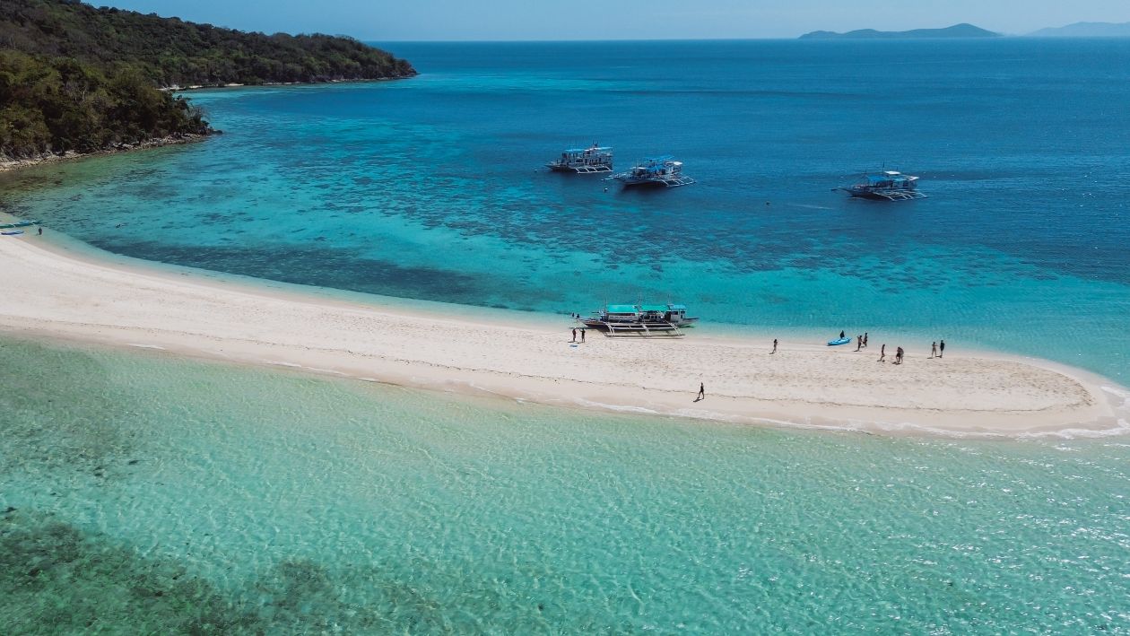 A stunning aerial view of a sandbar in the Philippines, surrounded by turquoise waters with traditional boats anchored nearby and people enjoying the beach, highlighting the affordability of island hopping and the low Philippines travel cost.