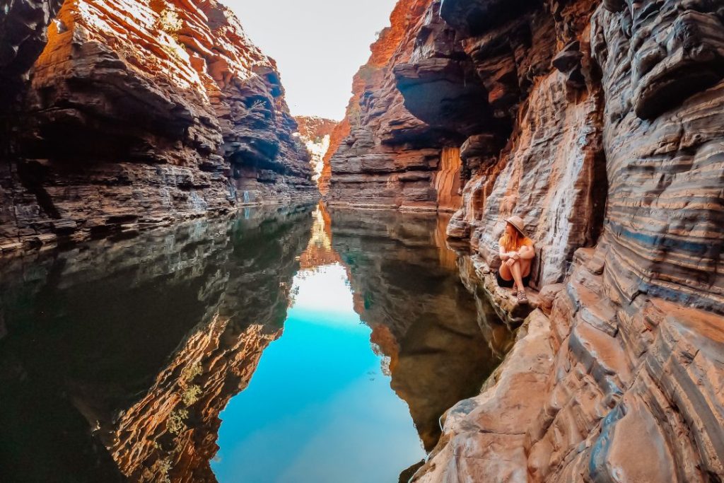 A woman in a hat sits on the rock ledge beside a tranquil waterhole, surrounded by steep, layered rock walls in one of the best gorges in Karijini National Park, Western Australia. The calm water reflects the red cliffs and bright sky.