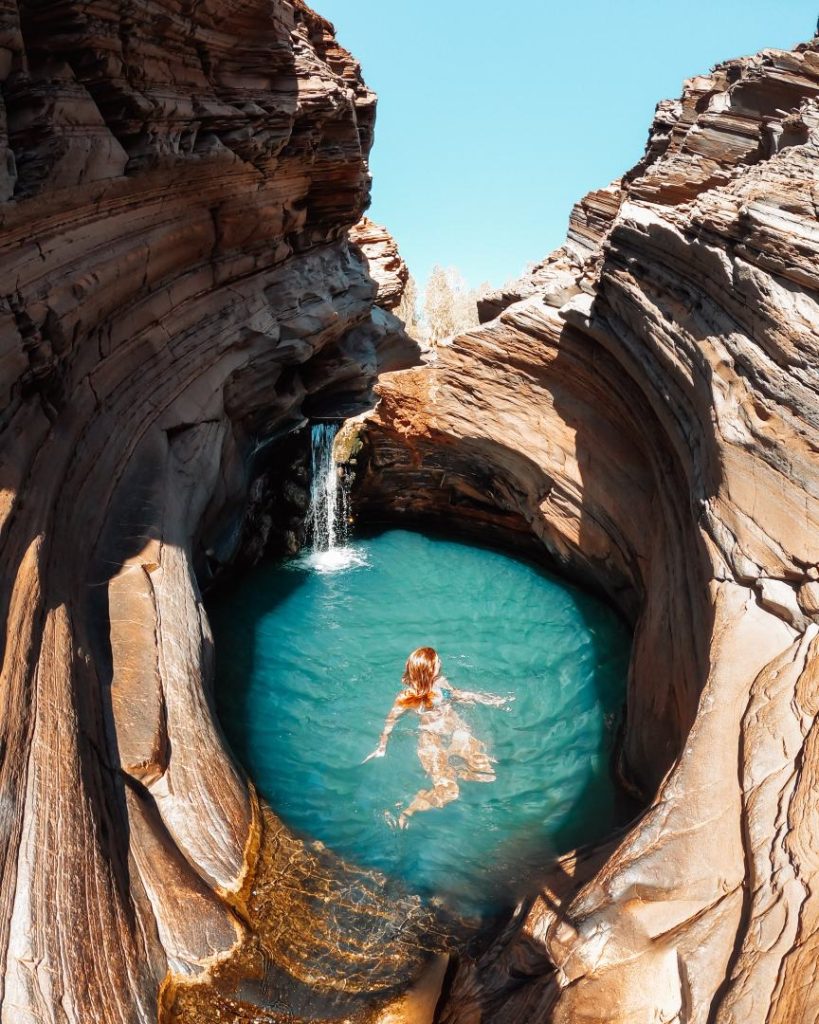 A woman swims in the turquoise rock pool of Hamersley Gorge, framed by smooth, curved red rock walls under a bright sky—one of the best times to visit Karijini for swimming and scenic beauty.