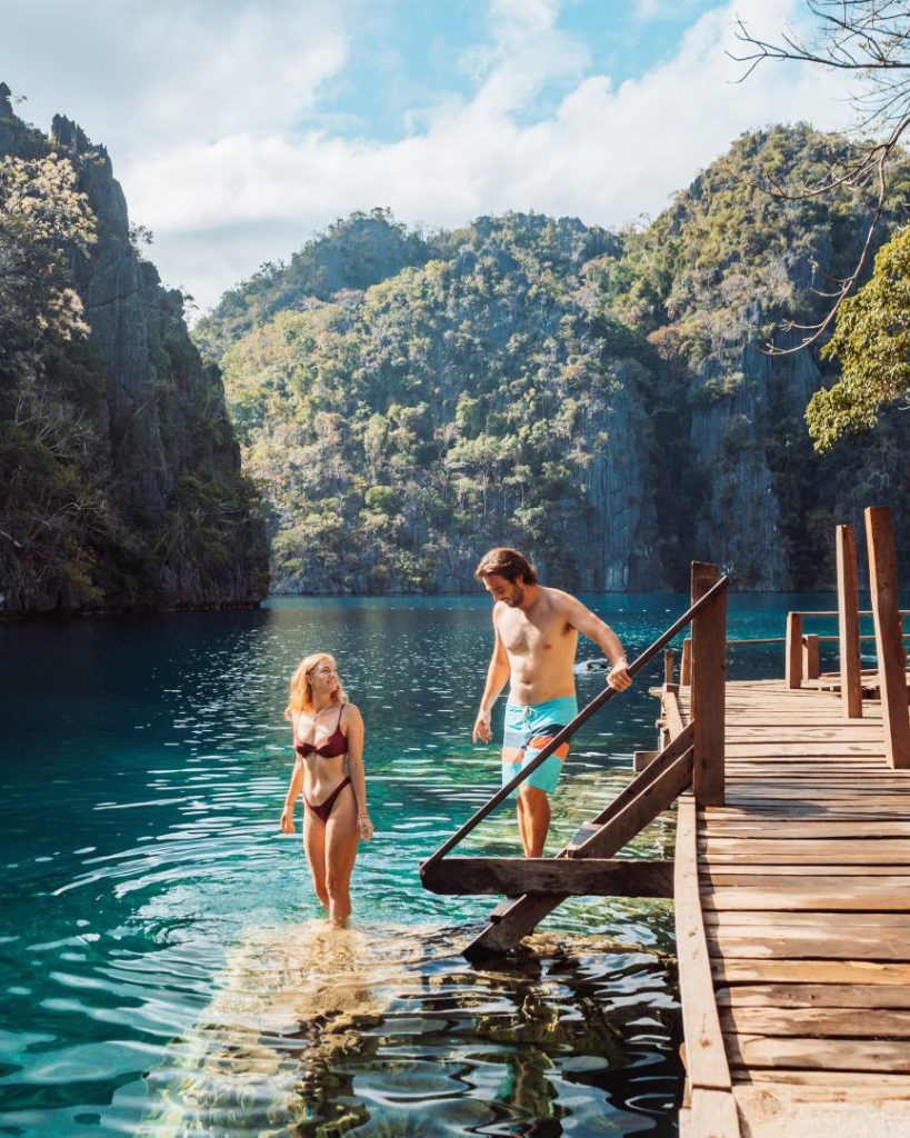 A couple enjoying the clear waters of Kayangan Lake in Coron, Philippines, stepping into the lake from a wooden dock, surrounded by lush limestone cliffs, showcasing the budget-friendly cost of traveling the Philippines.