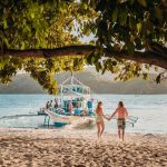 A couple walking hand-in-hand along a sandy beach towards a traditional Filipino boat, emphasizing how island hopping is an affordable part of the Philippines travel cost experience.