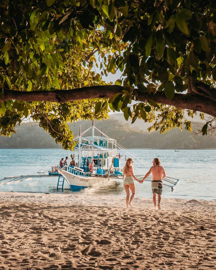 A couple walking hand-in-hand along a sandy beach towards a traditional Filipino boat, emphasizing how island hopping is an affordable part of the Philippines travel cost experience.