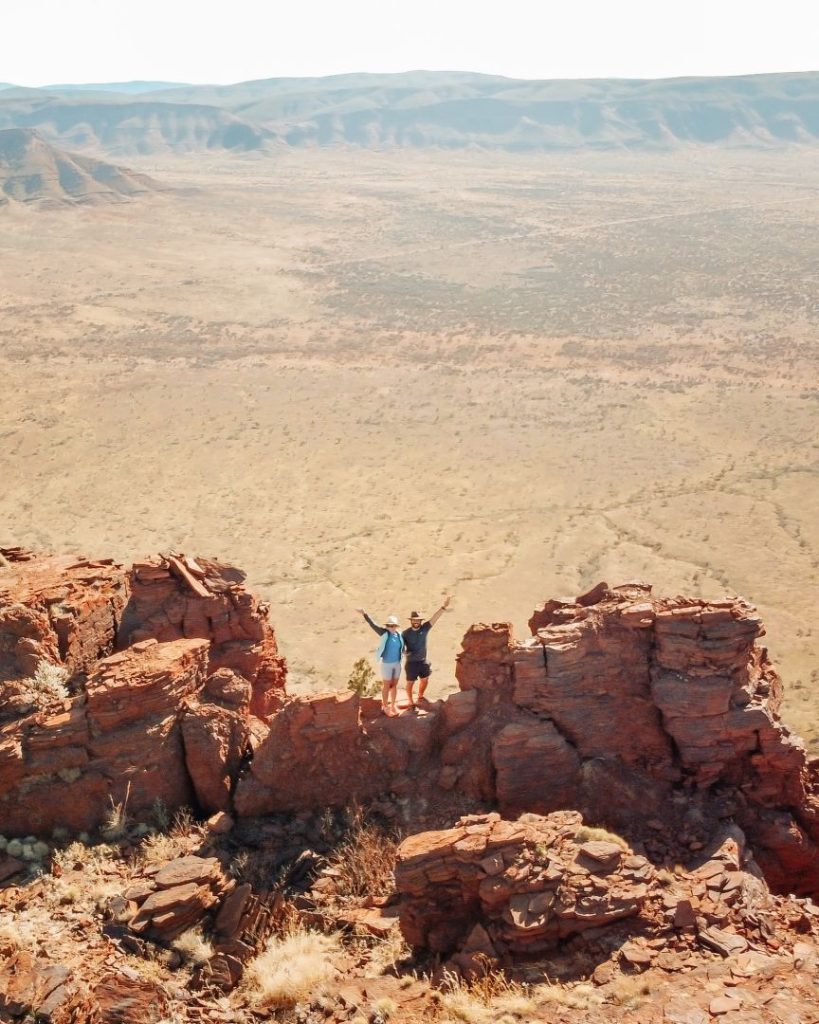Two hikers stand triumphantly on red rocky cliffs overlooking the vast outback plains from Mount Bruce, an epic Karijini National Park view during the best time to visit Karijini for hiking, winter.