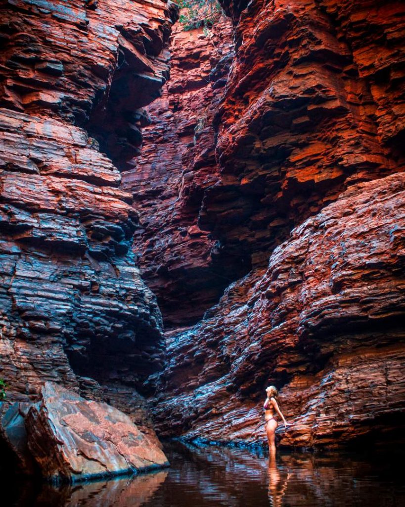 A woman stands waist-deep in the calm waters of Handrail Pool, surrounded by towering rust-red gorge walls—perfect conditions highlighting the best time to visit Karijini.