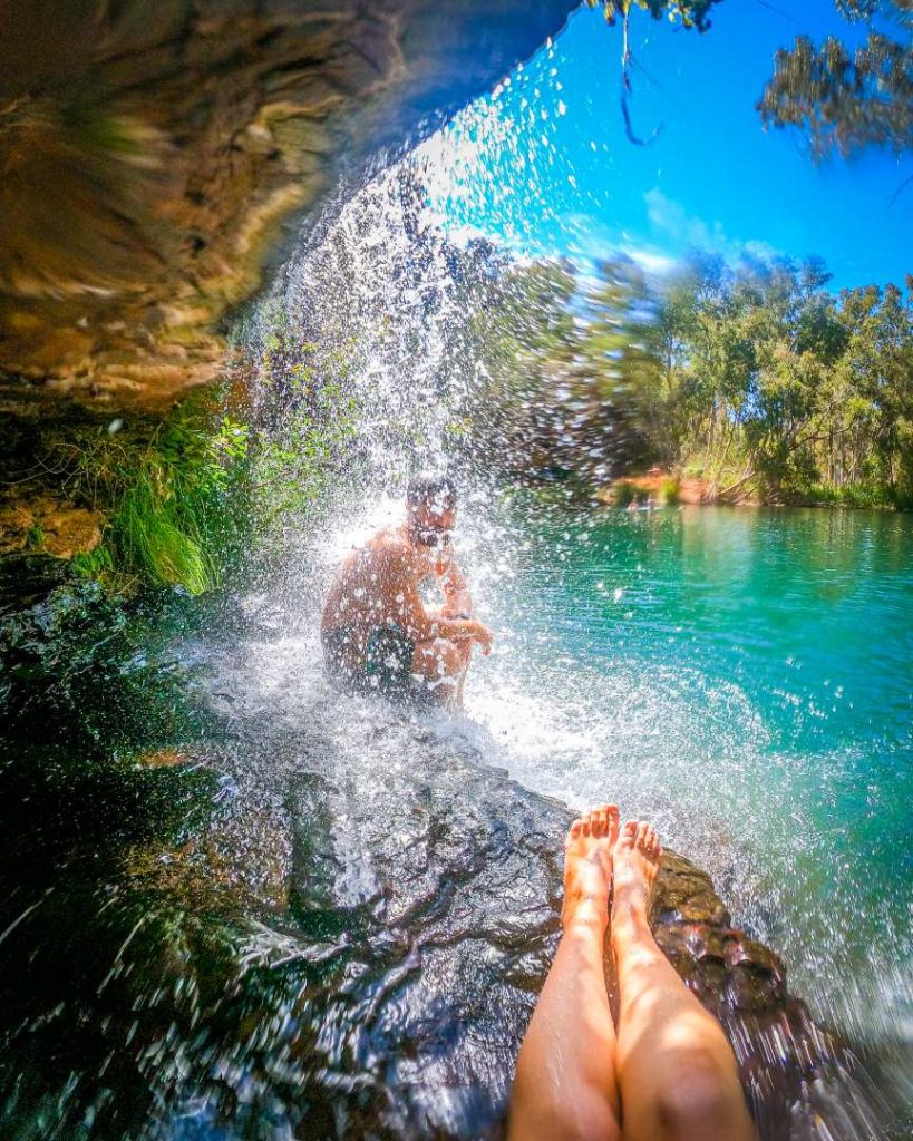 A man sits behind a waterfall at Fern Pool while another person’s legs rest in the foreground, capturing a refreshing moment during the best time to visit Karijini National Park.