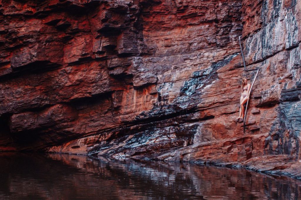 A woman climbs a metal handrail embedded in the cliffside above a still gorge pool, capturing the thrill of adventure during the best time to visit Karijini.