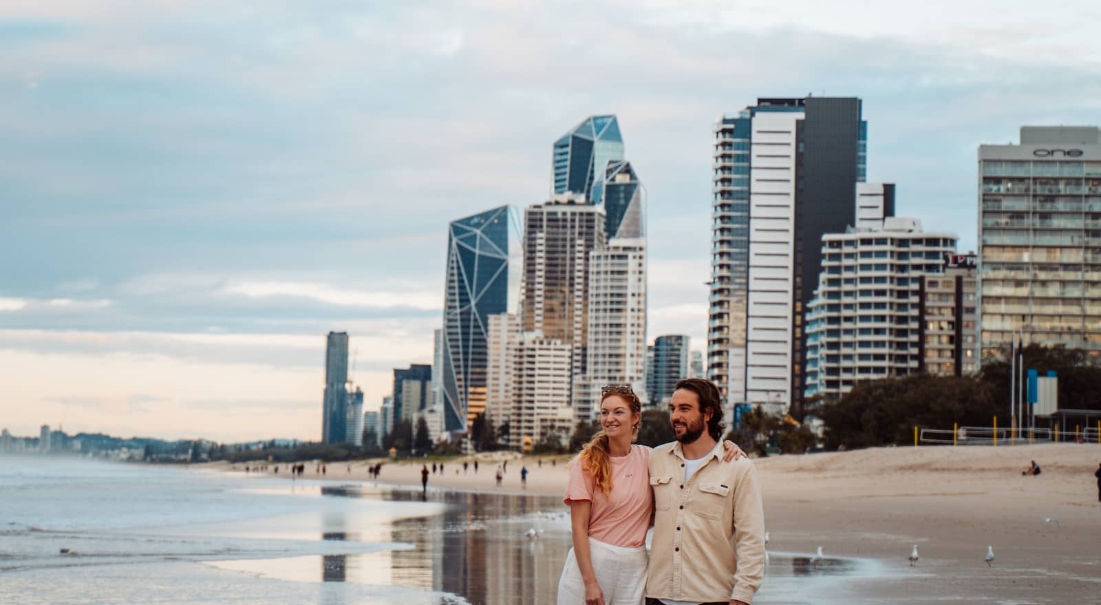 A couple stands on the wet sand of a Gold Coast beach, with the city's modern skyline towering behind them. The sky is overcast with soft, diffused light, and reflections of the buildings shimmer on the shoreline. A few beachgoers and seagulls are visible in the distance, adding to the serene coastal atmosphere.
