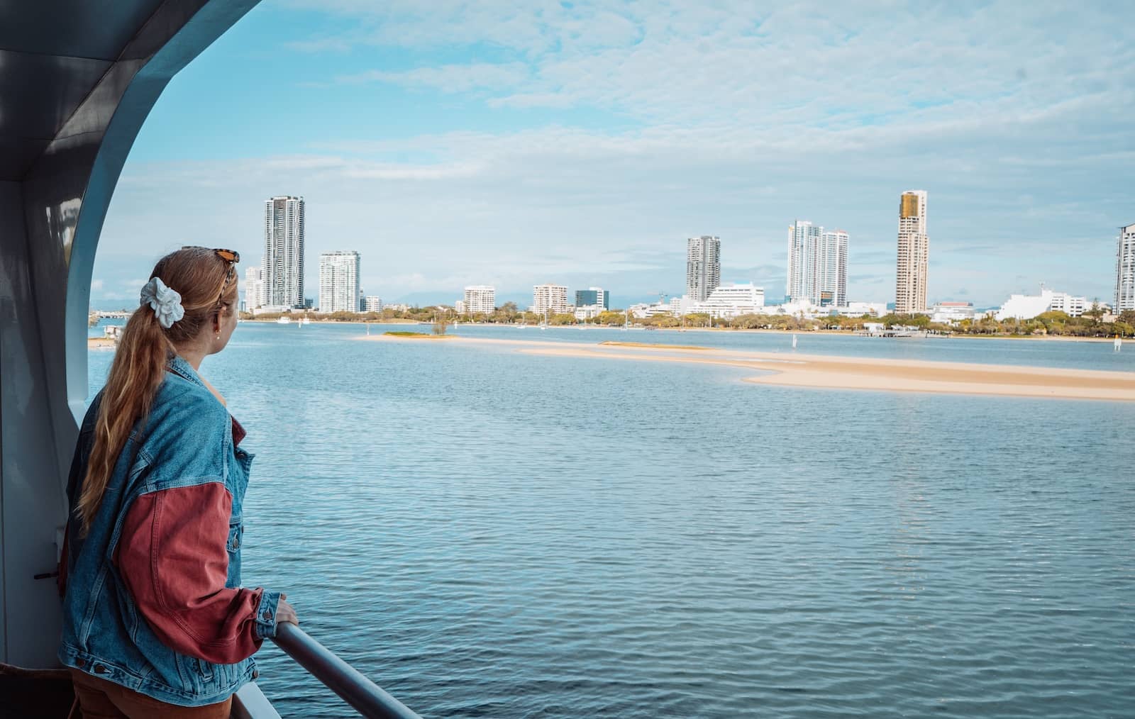 A woman wearing a denim jacket and a white hair scrunchie gazes out over the calm waters during a humpback whale cruise in Gold Coast, Australia. The skyline of high-rise buildings stretches across the horizon, contrasting with the blue sky and sandy patches along the water. The peaceful scene captures a blend of urban and coastal beauty.