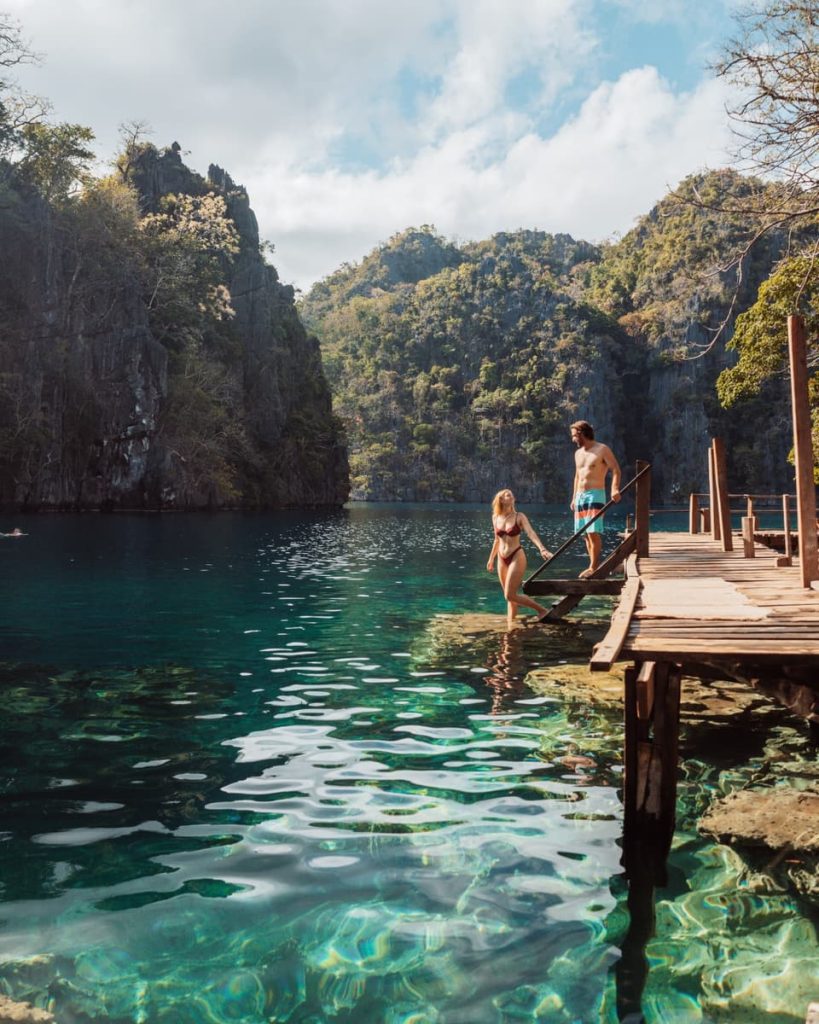 A couple walks down a wooden jetty into the clear water of Kayangan Lake, surrounded by lush limestone cliffs—a highlight of the best tours in Coron.