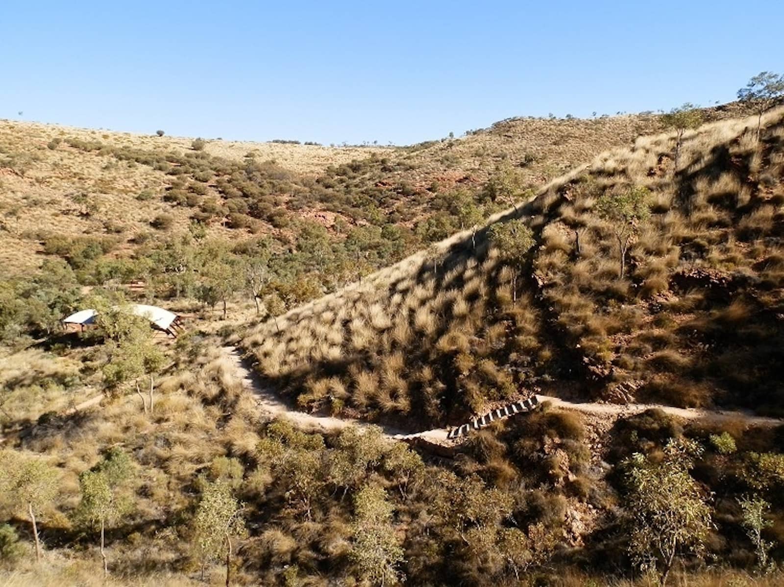 A winding dirt trail cuts through dry grassy hills with scattered trees, leading to a small footbridge and shaded shelters nestled in the Australian outback.