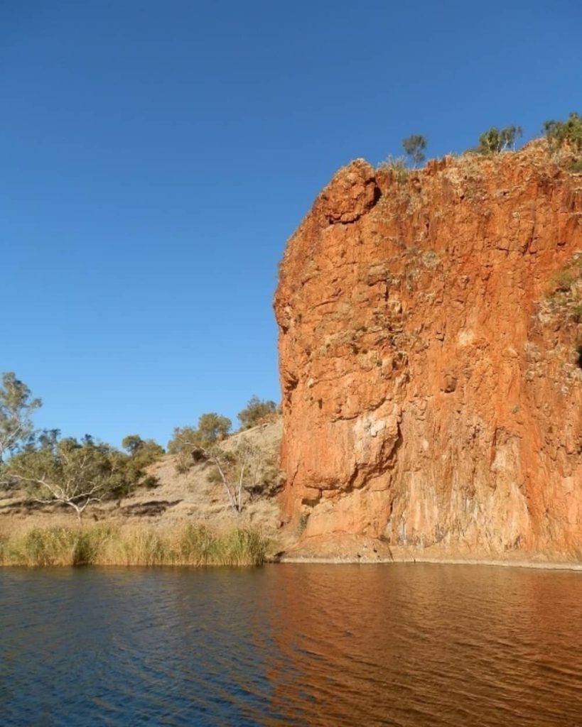 A towering red rock cliff rises dramatically from the edge of a calm waterhole, surrounded by dry grass, sparse trees, and a clear blue sky in the Glen Helen Gorge area.