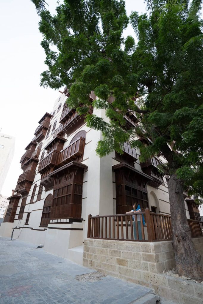 A woman in a long white skirt and blue scarf walks along a narrow alleyway in Jeddah's Al-Balad, framed by historic buildings with intricate wooden balconies—a lovely thing to do in Jeddah