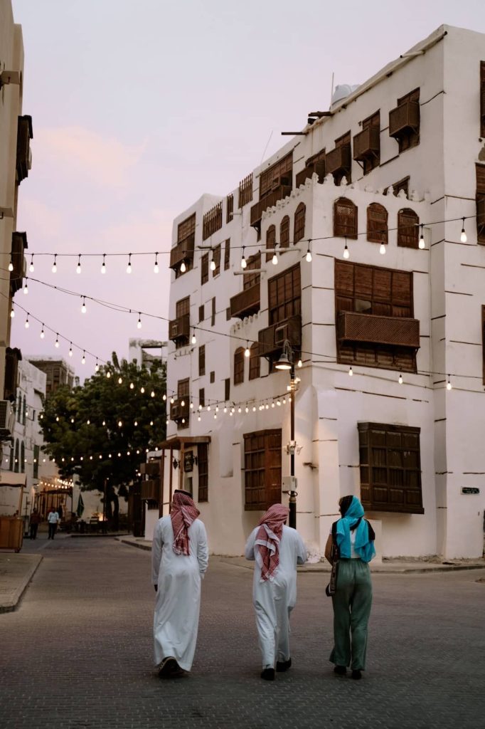 Three people walk under string lights in Al-Balad, Jeddah’s historic district, surrounded by whitewashed buildings with wooden window screens—exploring this area is a must-do thing in Jeddah for culture lovers.