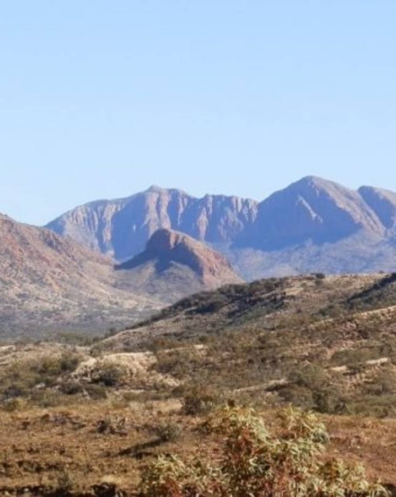 A panoramic view of the rugged West MacDonnell Ranges with Mt Sonder standing prominently in the background under a clear blue sky. The foreground features rolling, arid terrain dotted with dry shrubs and rocky outcrops.