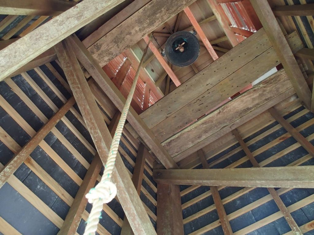 View looking up inside the wooden bell tower of St. Barnabas Church on Norfolk Island, showing the central bell hanging from beams and a thick rope extending downward.