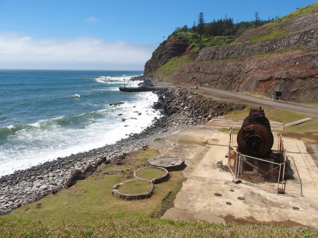 Rusted machinery from an old whaling station sits on a concrete platform above the rocky coastline of Cascade Bay, Norfolk Island, with the Cascade Jetty extending into the ocean and dramatic red cliffs rising in the background.