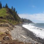 Rocky coastline at Cascade Bay, Norfolk Island, with waves crashing along the shore and an old rusted piece of machinery from a whaling station in the foreground. Green cliffs rise steeply in the background, dotted with tall Norfolk pine trees under a bright blue sky.