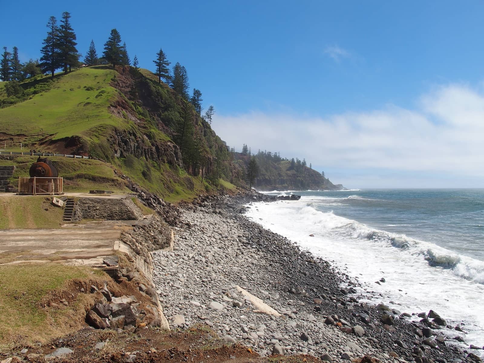 Rocky coastline at Cascade Bay, Norfolk Island, with waves crashing along the shore and an old rusted piece of machinery from a whaling station in the foreground. Green cliffs rise steeply in the background, dotted with tall Norfolk pine trees under a bright blue sky.