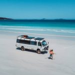 A couple celebrates by a white off-road campervan parked on the white sands of Cape Le Grand Beach, with turquoise water and deep blue ocean in the background