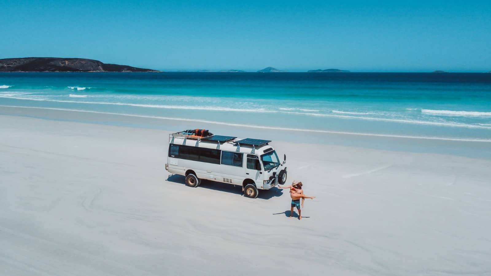 A couple celebrates by a white off-road campervan parked on the white sands of Cape Le Grand Beach, with turquoise water and deep blue ocean in the background
