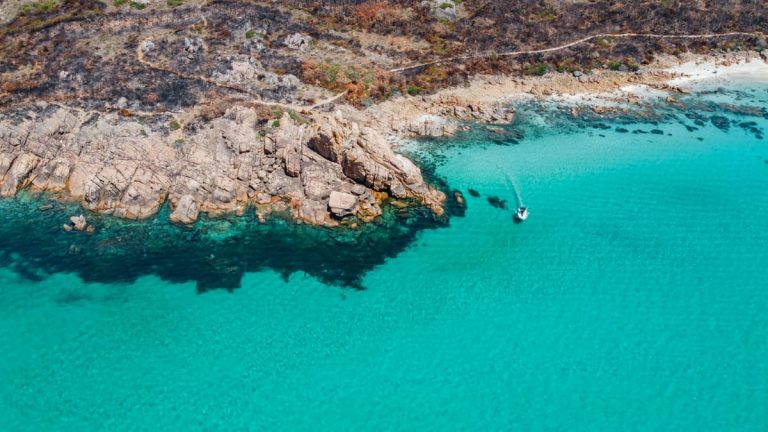 Aerial view of a small boat cruising along vibrant turquoise waters near rocky cliffs on the Perth to Margaret River road trip. The clear sea contrasts with the rugged shoreline and sparse coastal vegetation.