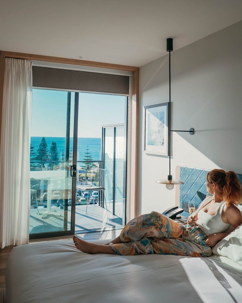 Woman sitting on a bed with white sheets overlooking the ocean from a window in the Hilton Inn Busselton, Western Australia