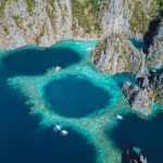 Aerial view of the Twin Lagoon in Coron, Palawan, with turquoise waters, dark blue pools, and dramatic limestone cliffs rising from the sea. Traditional Filipino boats float nearby.