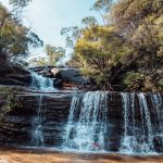 A wide-angle view of a tiered waterfall cascading over dark rock ledges, surrounded by bushland in the Blue Mountains region of NSW.