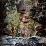 Woman in a red swimsuit standing on wet rock ledge above the secret infinity pool in Leura Cascades, Blue Mountains, surrounded by steep sandstone cliffs and dense green bushland.